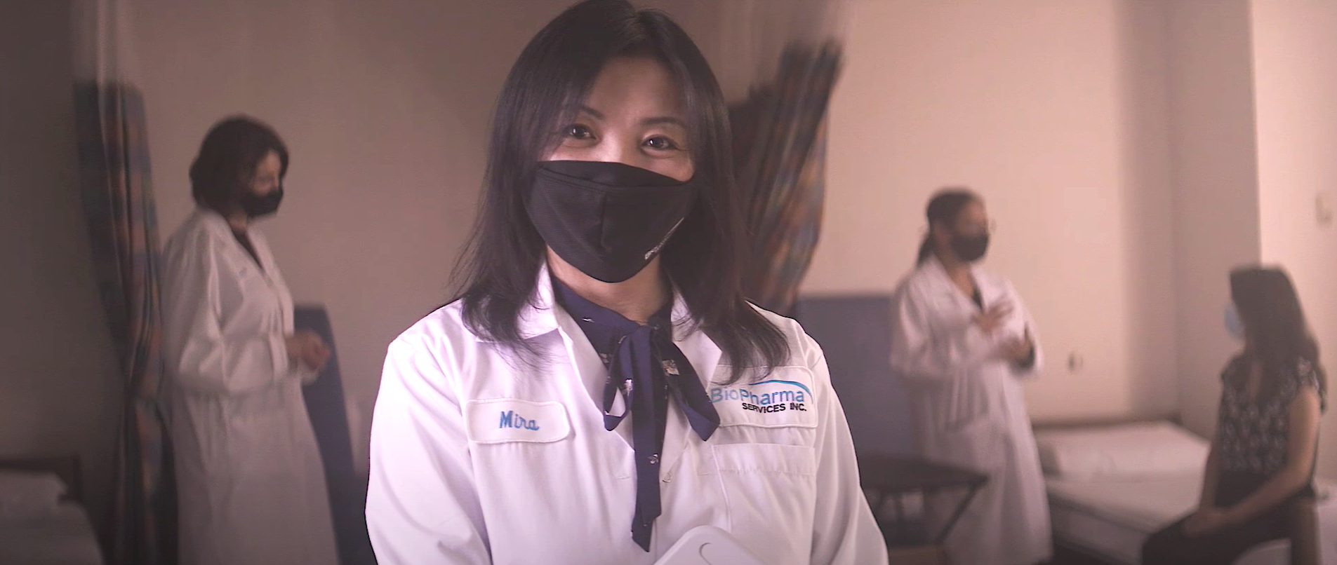A clinical study team member looks at the camera while volunteers are being attended to in the background.