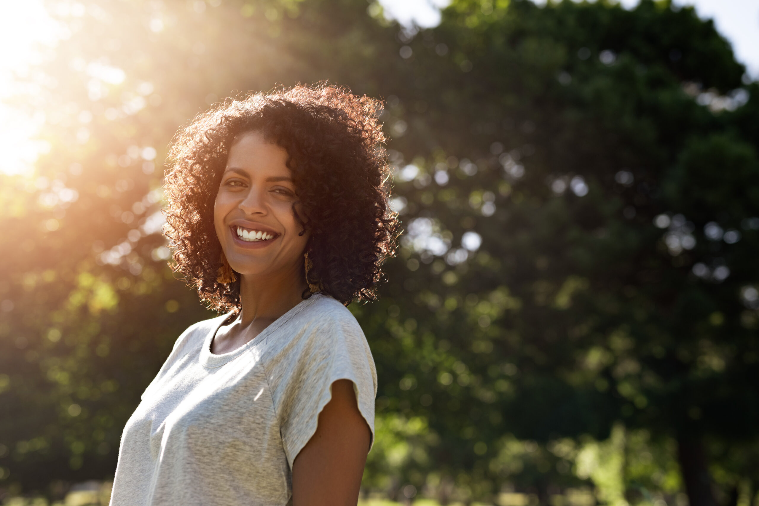 Portrait of a young woman with curly hair smiling while standing outside in a park on a sunny summer afternoon